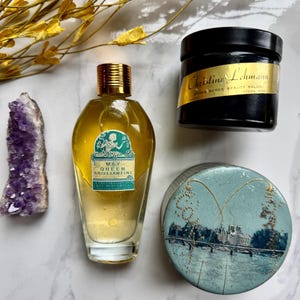 May include: An assortment of vintage apothecary items, including a bottle of May Queen Brilliantine, a black jar with gold lettering, a round tin, and a purple amethyst crystal. The items are arranged on a marble surface with dried yellow foliage.