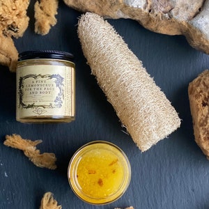 May include: A fine lemonsrub for the face and body, a jar of yellow scrub, a loofah, and natural sponges are arranged on a dark surface. The jar has a black lid and a label with text. The scrub is in an open glass jar.