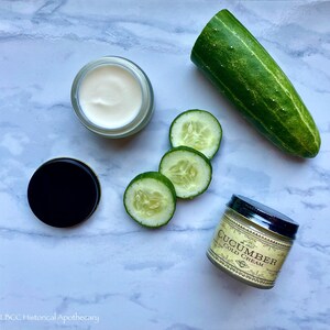 May include: A white jar of cucumber cold cream with a black lid, next to a jar of cucumber cold cream with a gold lid and a label that reads "Cucumber Cold Cream". There are also four slices of cucumber on a white marble surface.