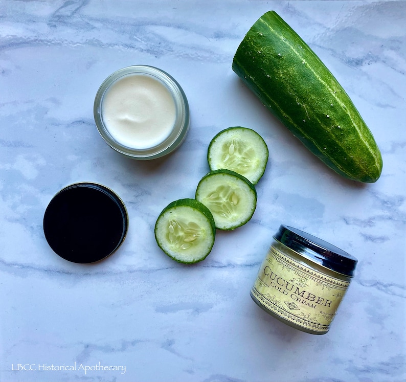 May include: A white jar of cucumber cold cream with a black lid, next to a jar of cucumber cold cream with a gold lid and a label that reads "Cucumber Cold Cream". There are also four slices of cucumber on a white marble surface.