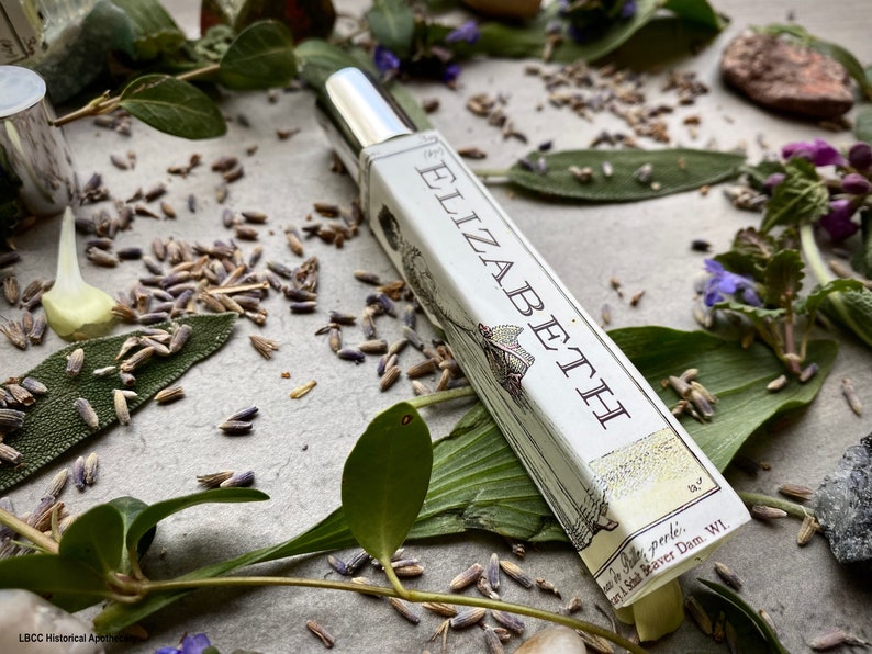 May include: A silver and white bottle of perfume with the word "Elizabeth" printed on the label. The bottle is surrounded by green leaves and lavender flowers.