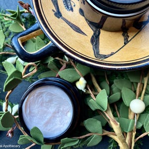 May include: A close-up of a small jar of cream with a black lid. The jar is sitting on a table with a green leafy branch and a ceramic plate with a black and gold design. The text "LBCC Historical Apothecary" is visible at the bottom of the image.