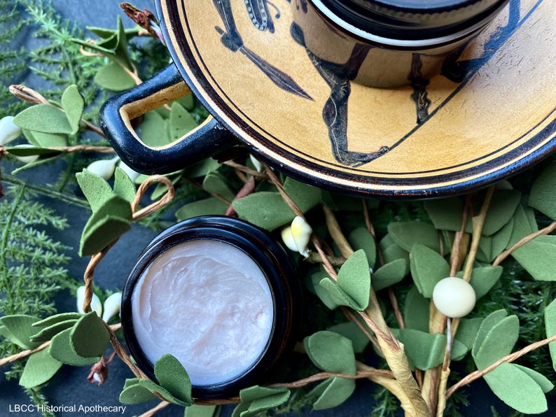 May include: A close-up of a small jar of cream with a black lid. The jar is sitting on a table with a green leafy branch and a ceramic plate with a black and gold design. The text "LBCC Historical Apothecary" is visible at the bottom of the image.