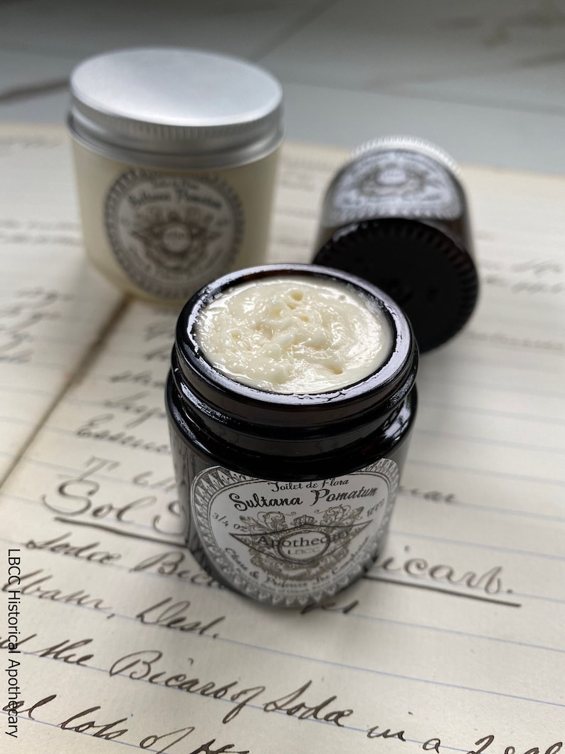 May include: A small glass jar with a black lid and a white label that reads "Joilet de Flora Sultana Pomatum 3/4 oz." The jar is filled with a white cream and is sitting on a piece of paper with handwritten text.