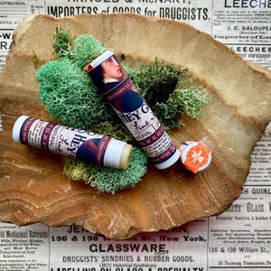May include: Two tubes of lip balm with brown and white labels. The labels say "Hey Girl" and "Fine & Natural". The tubes are sitting on a piece of brown rock with green moss.