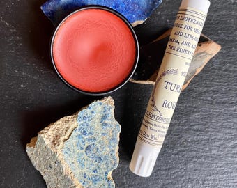 May include: A small, round, black-rimmed container filled with red rouge sits next to a tube of lipstick. The lipstick tube is labeled "Ture Rouge" and "Historical Apothecary." Fragments of blue and beige stone surround the products.
