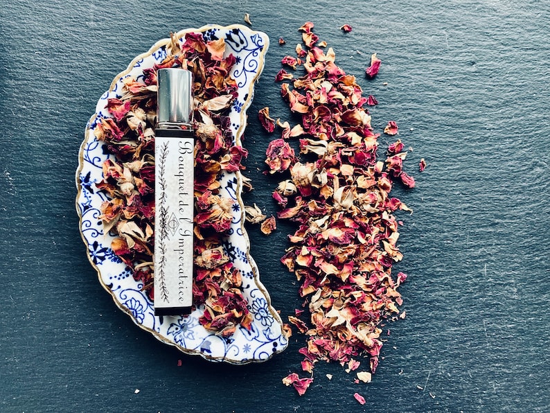 K&ouml;nnte beinhalten: Eine Glasflasche mit Parf&uuml;m, einem silbernen Verschluss und einem wei&szlig;en Etikett mit der Aufschrift "Bouquet de l'Imp&eacute;ratrice". Die Flasche steht auf einer blauen und wei&szlig;en Porzellanschale mit getrockneten Rosenbl&auml;ttern. Die Schale ist halbmondf&ouml;rmig. Getrocknete Rosenbl&auml;tter sind um die Schale herum verstreut.