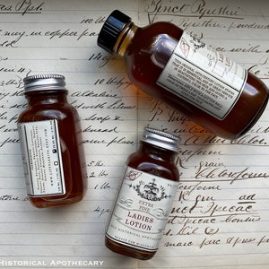 May include: Three small glass bottles filled with a brown liquid. The bottles have silver lids and labels with text. The labels are white with black text. The bottles are sitting on a piece of paper with handwritten text. The text on the labels reads "Extra Fine Ladies Lotion" and "This original 19th Century Lotion states that it is beneficial and of great service in beautifying the skin and giving a youthful color to the face."