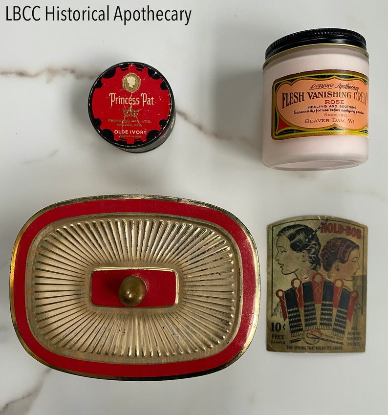 May include: A collection of vintage apothecary items, including a Princess Pat tin, a jar of Flesh Vanishing Cream, a decorative gold and red container, and a Hold-Bob sample card. The items are arranged on a light-colored surface.