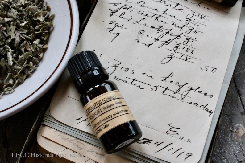 May include: A small brown glass bottle with a black cap, labeled "Eucalyptus (Globulus) Wisconsin" and "with soft woody undertones". The bottle is sitting on a vintage notebook with handwritten text, including "Ziss in kaef glass water 3 times a day" and "E. M.D. 4/1/19".