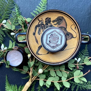 May include: A small tin of Parisian Pomade for dressing the hair, sitting on a decorative plate with a black and gold design. The plate features a scene of a man with a bow and arrow and a lion. The tin is surrounded by green foliage.