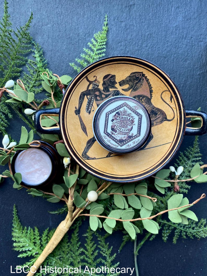 May include: A small tin of Parisian Pomade for dressing the hair, sitting on a decorative plate with a black and gold design. The plate features a scene of a man with a bow and arrow and a lion. The tin is surrounded by green foliage.