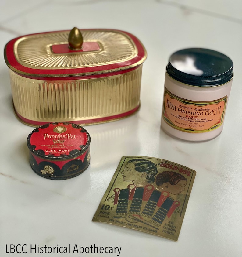 May include: An assortment of vintage apothecary items, including a gold and red container, a pink jar of "Flesh Vanishing Cream," and a small tin of "Princess Pat" powder. A "Hold-Bob" hair accessory card is also present.