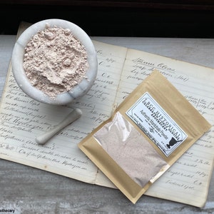 May include: A brown paper bag with a label that reads "LBCC Historical Apothecary Authentic Maréchale Powder". The bag is filled with a light brown powder. The bag is sitting on top of an open book with handwritten notes.