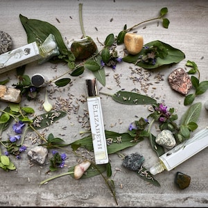 May include: Three glass roller bottles with labels that say "Elizabeth" and "LBCC Historical Apothecary". The bottles are surrounded by green leaves, purple flowers, and various stones.
