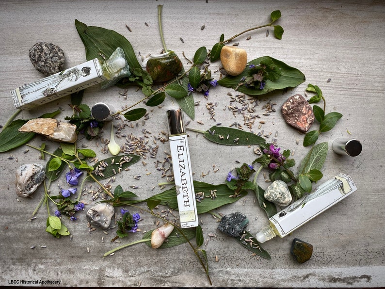 May include: Three glass roller bottles with labels that say "Elizabeth" and "LBCC Historical Apothecary". The bottles are surrounded by green leaves, purple flowers, and various stones.