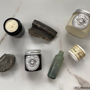 May include: A collection of apothecary items on a white marble surface. The items include a small glass bottle, a small tin box, a small jar of cream, and a larger jar of cream. The jars of cream are labeled "Saliana Pamaton" and "Hand Lotion".