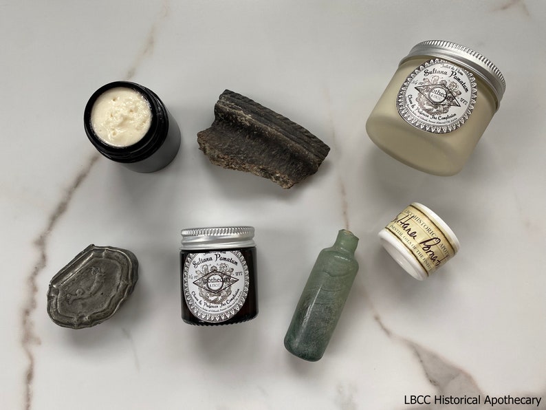 May include: A collection of apothecary items on a white marble surface. The items include a small glass bottle, a small tin box, a small jar of cream, and a larger jar of cream. The jars of cream are labeled "Saliana Pamaton" and "Hand Lotion".