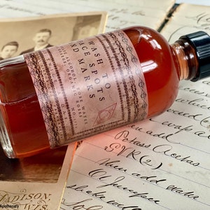 May include: A glass bottle with a brown liquid and a black cap. The label reads "Wash To Face Spots And Marks" with the text "Rose Water, Alcohol, Organic Roses" below.