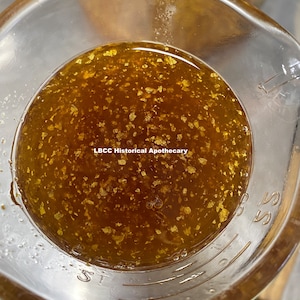 May include: Close-up of a clear glass measuring cup filled with a golden liquid and gold flakes. The text "LBCC Historical Apothecary" is visible. The cup has measurement markings.