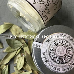K&ouml;nnte beinhalten: Ein kleines Glasgef&auml;&szlig; mit einem wei&szlig;en Etikett, auf dem "LBCC Apothecary Golden Ointment Cold Soothing" steht, und eine silberne Metalldose mit einem wei&szlig;en Etikett, auf dem "LBCC Historical Apothecary Cold Cream Beaver Dam" steht.
