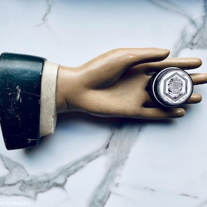 May include: A small, round, silver tin with a black label and white text that reads "Lavender & Eucalyptus" on a white background. The tin is held in a plaster hand.