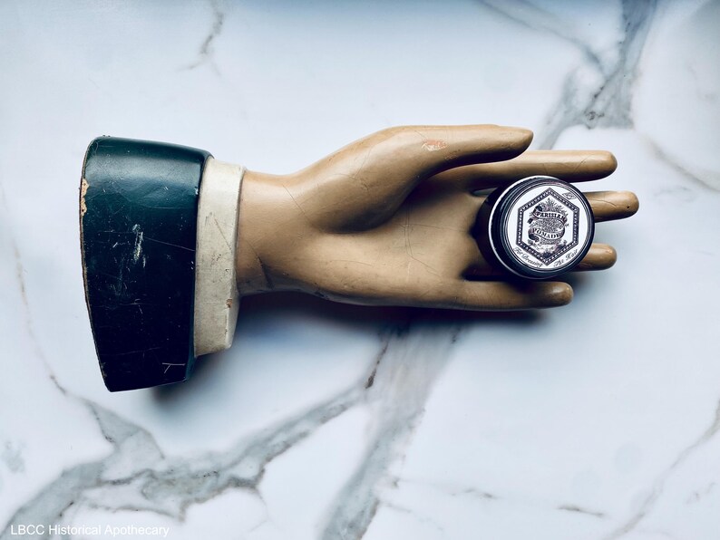 May include: A small, round, silver tin with a black label and white text that reads "Lavender & Eucalyptus" on a white background. The tin is held in a plaster hand.