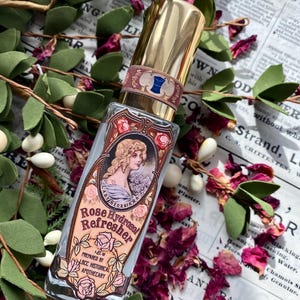 K&ouml;nnte beinhalten: Eine durchsichtige Glasflasche mit goldener Kappe und einem Etikett mit der Aufschrift "Rose Hydrosol Refresher 1 oz. Prepared by LBCC Historical Apothecary". Das Etikett zeigt eine Vintage-Illustration einer Frau mit Blumen und einem blauen Band.