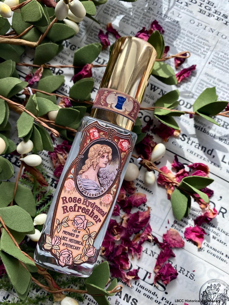 K&ouml;nnte beinhalten: Eine durchsichtige Glasflasche mit goldener Kappe und einem Etikett mit der Aufschrift "Rose Hydrosol Refresher 1 oz. Prepared by LBCC Historical Apothecary". Das Etikett zeigt eine Vintage-Illustration einer Frau mit Blumen und einem blauen Band.