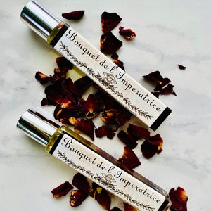 K&ouml;nnte beinhalten: Zwei silberne Rollflaschen mit einem wei&szlig;en Etikett, auf dem "Bouquet de l'Imp&eacute;ratrice" in schwarzer Schrift steht. Die Flaschen sind von roten Rosenbl&auml;ttern auf wei&szlig;em Hintergrund umgeben.