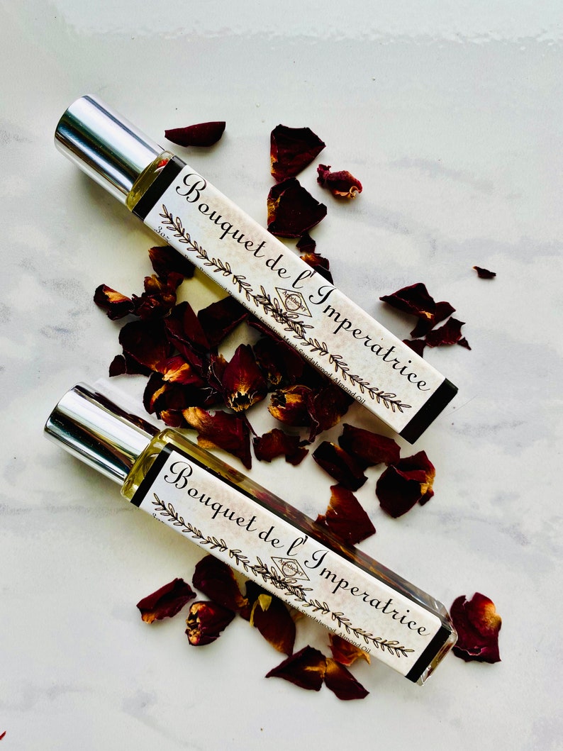 K&ouml;nnte beinhalten: Zwei silberne Rollflaschen mit einem wei&szlig;en Etikett, auf dem "Bouquet de l'Imp&eacute;ratrice" in schwarzer Schrift steht. Die Flaschen sind von roten Rosenbl&auml;ttern auf wei&szlig;em Hintergrund umgeben.