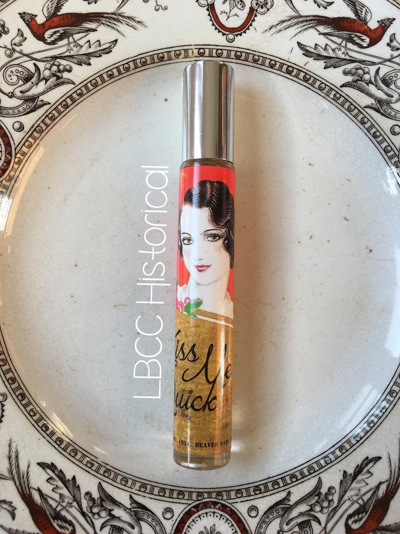 Peut inclure: Un flacon de parfum en argent avec un bouchon &agrave; bille. L'&eacute;tiquette est rouge et or et repr&eacute;sente une femme aux cheveux noirs portant une robe rouge. Le texte sur l'&eacute;tiquette est "Kiss Me Quick".