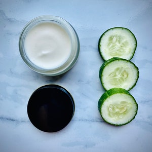 May include: A white cream in a glass jar with a black lid, next to three slices of green cucumber on a white marble surface.