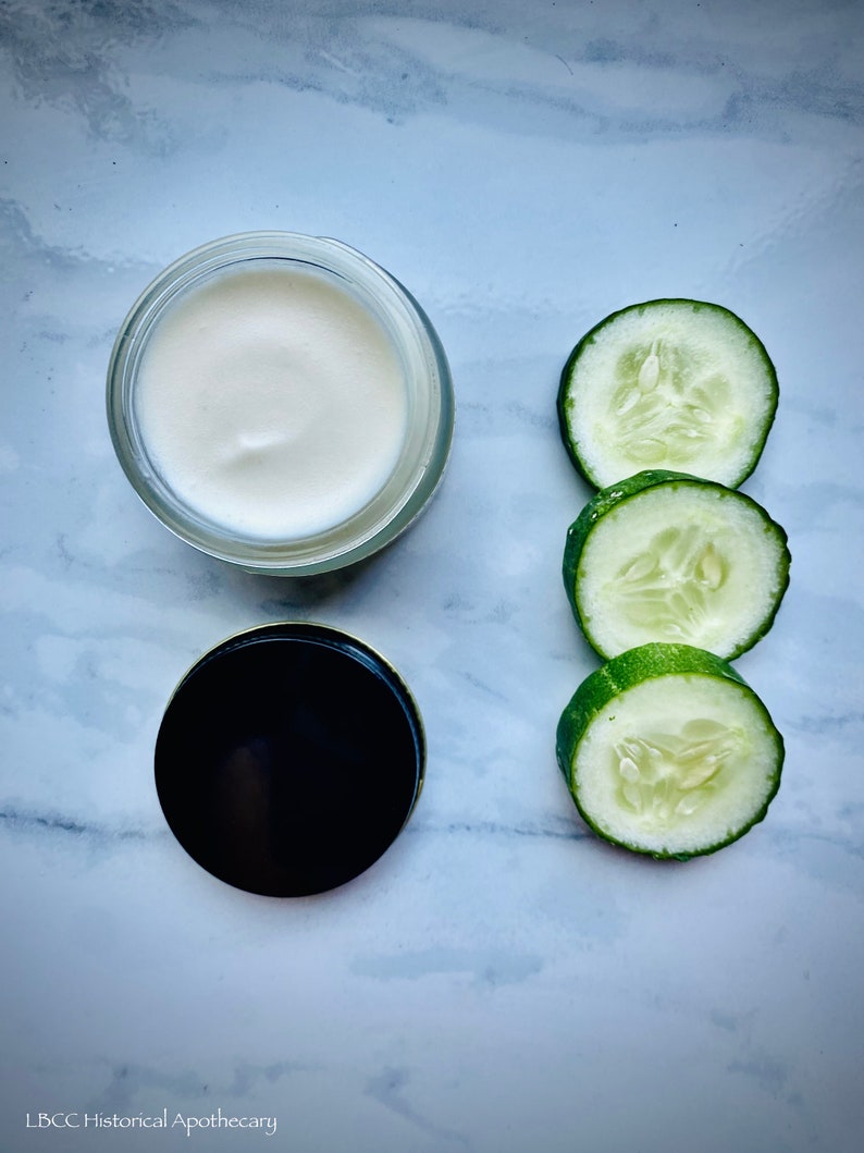 May include: A white cream in a glass jar with a black lid, next to three slices of green cucumber on a white marble surface.