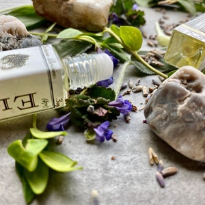 May include: A small glass bottle with a rollerball applicator filled with a clear liquid. The bottle is labeled "Botanical Perfumes" and "3 (1/4)". The bottle is surrounded by green leaves, purple flowers, and small gray stones.