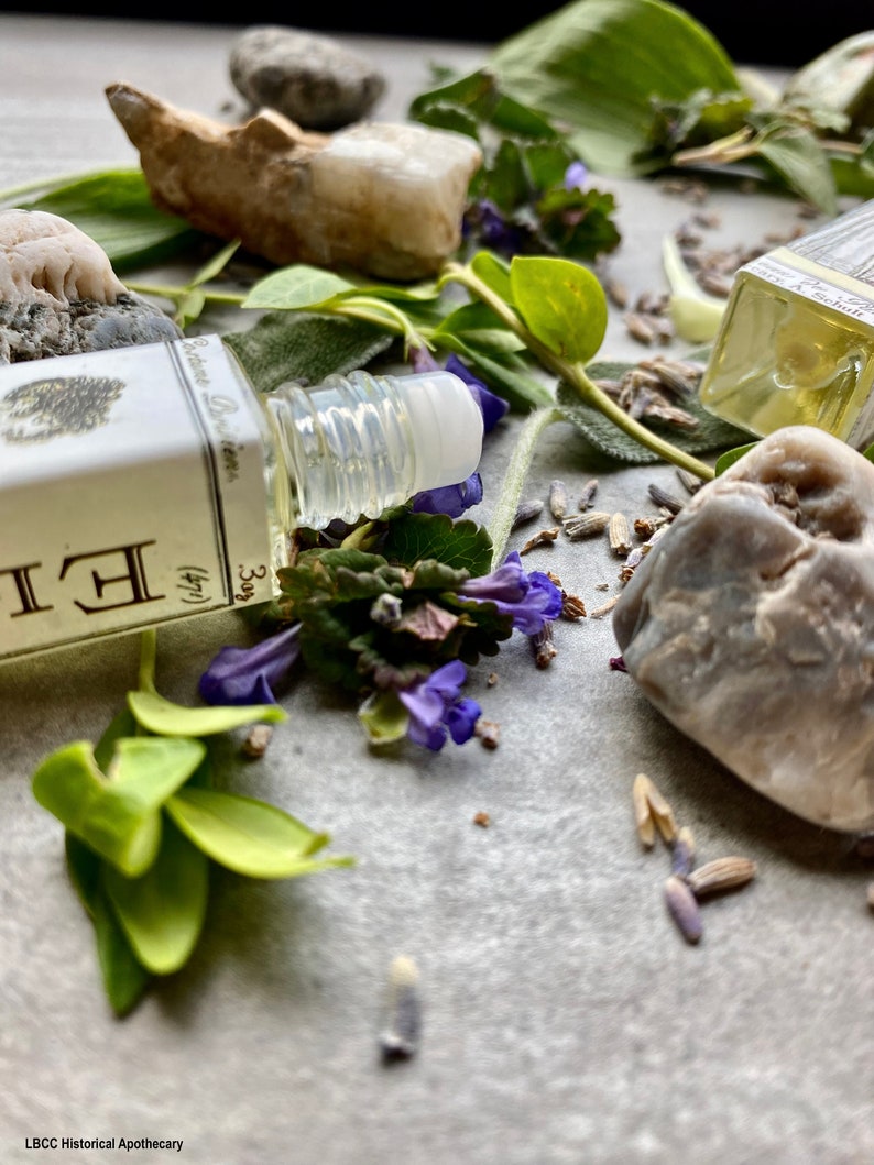 May include: A small glass bottle with a rollerball applicator filled with a clear liquid. The bottle is labeled "Botanical Perfumes" and "3 (1/4)". The bottle is surrounded by green leaves, purple flowers, and small gray stones.