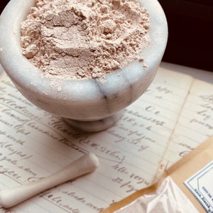 May include: A marble mortar filled with light brown powder sits on a vintage paper with handwritten text. A matching pestle rests nearby. A small, sealed packet of the powder is also visible. The image has a vintage apothecary theme.