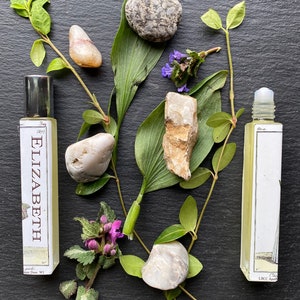May include: Two glass bottles of essential oil with labels that read "Elizabeth" and "Che LBCC Apo." The bottles are surrounded by green leaves, purple flowers, and grey stones.