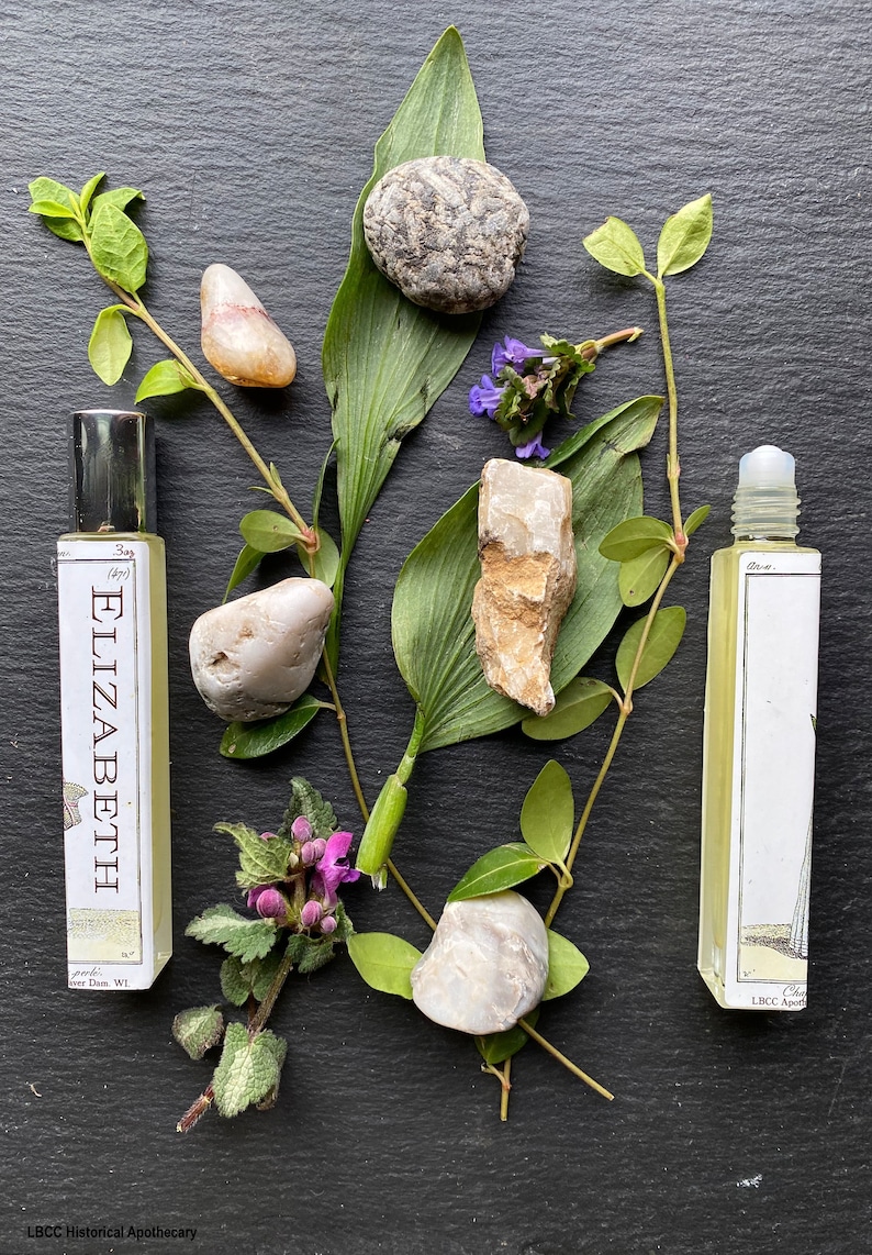 May include: Two glass bottles of essential oil with labels that read "Elizabeth" and "Che LBCC Apo." The bottles are surrounded by green leaves, purple flowers, and grey stones.