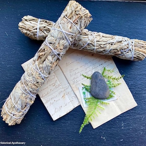 May include: Two white sage bundles tied with white string, resting on vintage paper with handwritten text. A gray stone and green sprigs are also on the paper. The background is a dark blue surface.