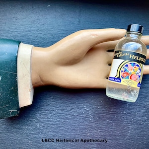 May include: A clear glass bottle with a black cap, containing a liquid. The bottle has a label with the text "Eau de Toilette HELIOTROPE" and a floral design. The bottle is held in a plaster hand.