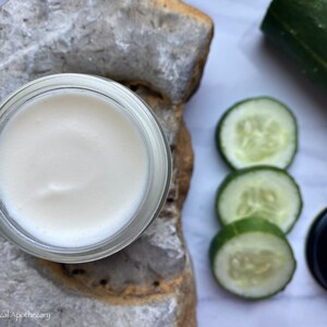 May include: A white cream in a glass jar with a lid. The jar is sitting on a piece of rough, light brown stone. There are three slices of cucumber on a white surface next to the jar.