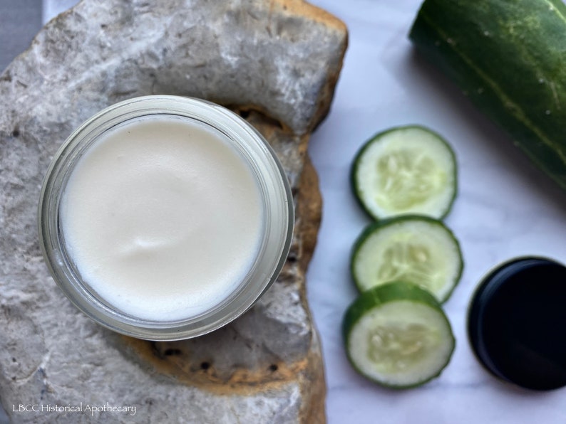 May include: A white cream in a glass jar with a lid. The jar is sitting on a piece of rough, light brown stone. There are three slices of cucumber on a white surface next to the jar.