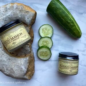 May include: Two jars of cucumber cold cream with a gold label and black lids. The jars are on a white marble surface with a cucumber and cucumber slices.