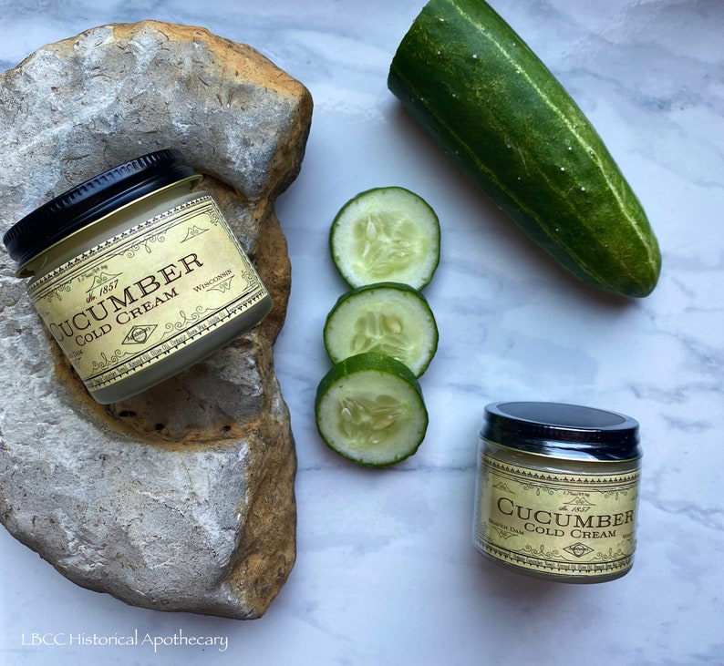 May include: Two jars of cucumber cold cream with a gold label and black lids. The jars are on a white marble surface with a cucumber and cucumber slices.