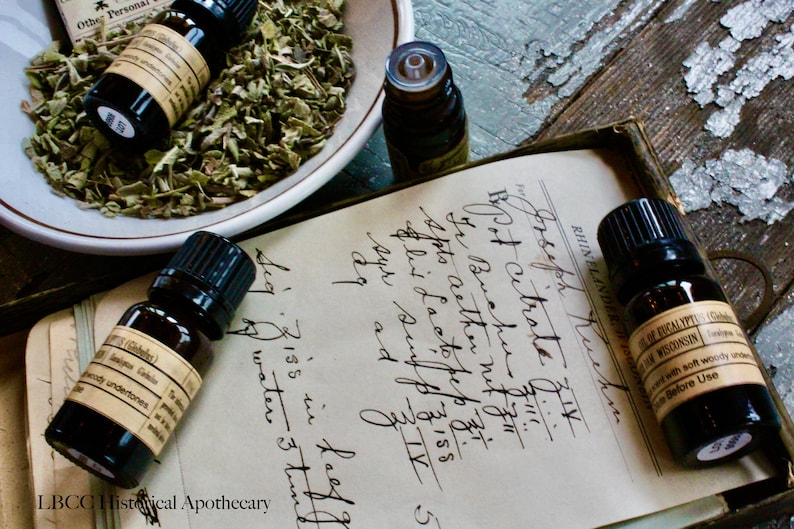May include: Four small brown glass bottles with black caps containing essential oils. The bottles are sitting on a vintage notebook with handwritten text. The text includes the words "Bpot citrate" and "Buchu".