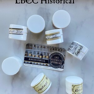K&ouml;nnte beinhalten: Eine Sammlung kleiner wei&szlig;er Gl&auml;ser mit Etiketten, auf denen "LBCC Historical Apothecary" und verschiedene D&uuml;fte stehen, darunter "Hastings", "Rose", "Enamel" und "Vapor Ice". Die Gl&auml;ser sind auf einer wei&szlig;en Marmorfl&auml;che angeordnet, auf der sich eine Visitenkarte mit dem Text "#LBCCHISTORICAL" befindet, die auch Social-Media-Handles enth&auml;lt. Der Text "Cream & Salve Samples" steht unter den Gl&auml;sern.
