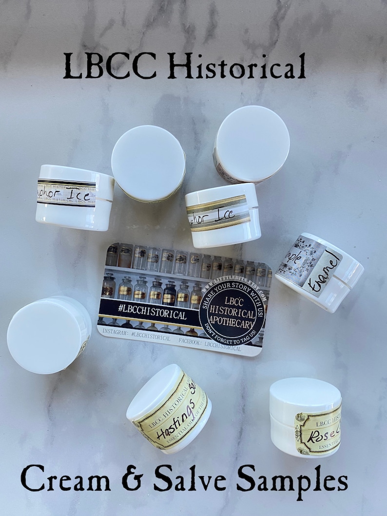 K&ouml;nnte beinhalten: Eine Sammlung kleiner wei&szlig;er Gl&auml;ser mit Etiketten, auf denen "LBCC Historical Apothecary" und verschiedene D&uuml;fte stehen, darunter "Hastings", "Rose", "Enamel" und "Vapor Ice". Die Gl&auml;ser sind auf einer wei&szlig;en Marmorfl&auml;che angeordnet, auf der sich eine Visitenkarte mit dem Text "#LBCCHISTORICAL" befindet, die auch Social-Media-Handles enth&auml;lt. Der Text "Cream & Salve Samples" steht unter den Gl&auml;sern.