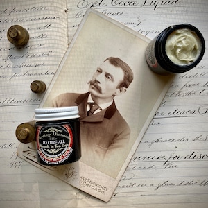 May include: A vintage notebook with handwritten notes and a black and white photograph of a man with a mustache. There are two small glass jars with labels, one labeled "Hastings Ointment" and the other labeled "Ext. Coca Liquid".