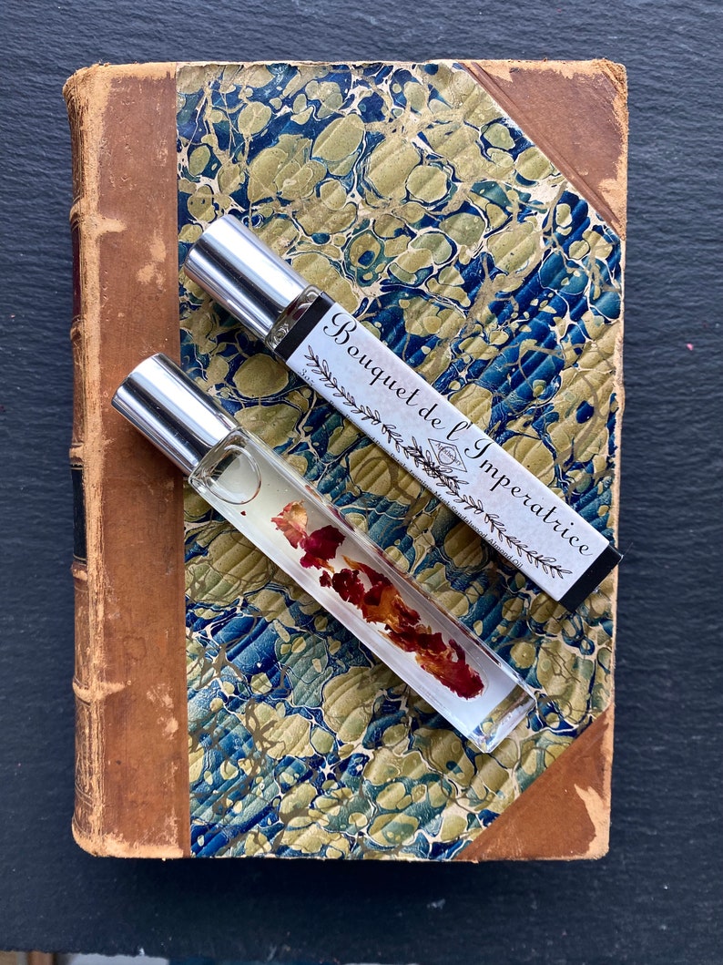 K&ouml;nnte beinhalten: Zwei transparente Glas-Rollflaschen mit silbernen Kappen und Etiketten. Das Etikett auf der oberen Flasche lautet "Bouquet de l'Imp&eacute;ratrice". Die Flaschen stehen auf einem alten Buch mit marmoriertem Einband.
